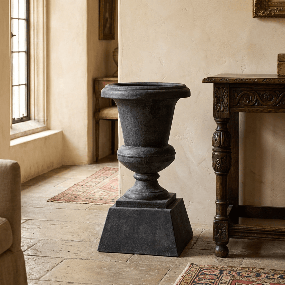 Hawthorne Aged Iron Urn Planter in Black, shown empty beside a carved wooden console in a traditional interior.