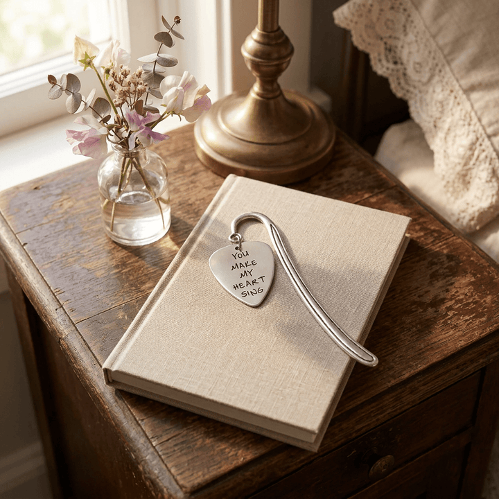 Silver-tone guitar-pick charm bookmark on a linen-covered journal beside a floral vase.