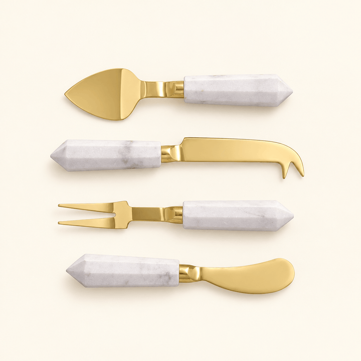 Four gold-finished cheese utensils with faceted white marble handles on an ivory background