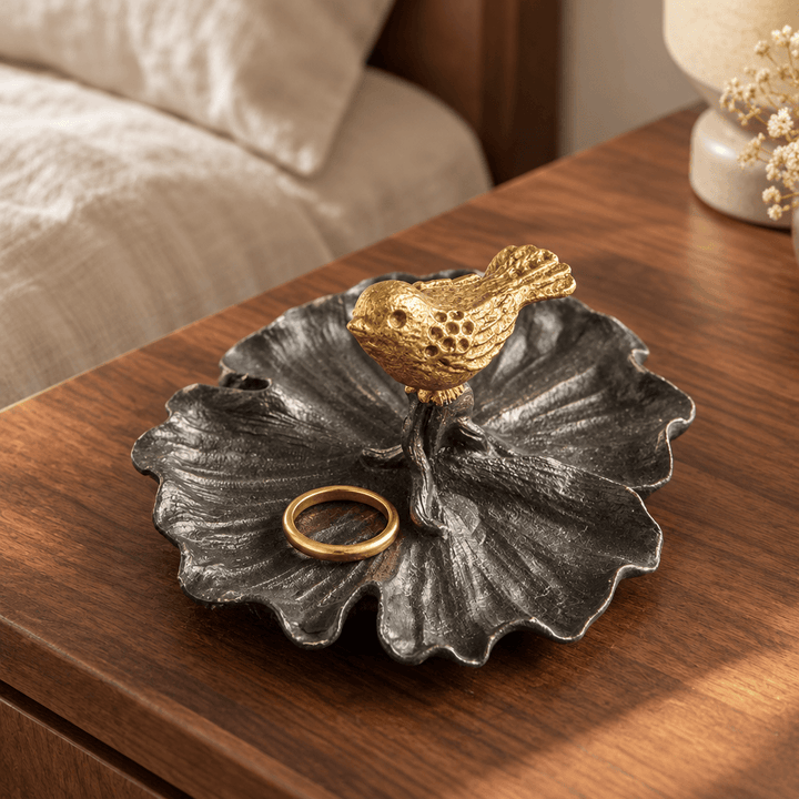 Pewter jewelry tray with a gold bird holding a ring on a walnut bedside table.