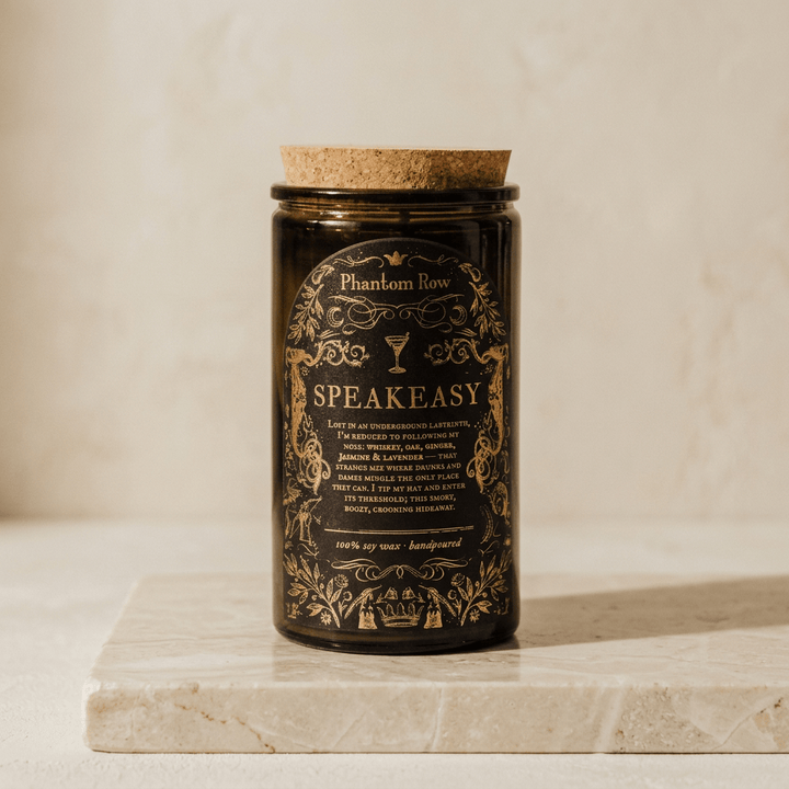 Tall Speakeasy candle in a dark glass jar with cork lid and ornate black-and-gold label on ivory stone.