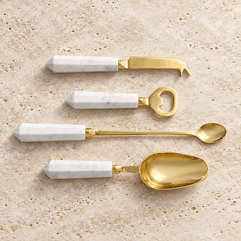 Four gold cocktail tools with faceted white marble handles displayed on pale travertine.
