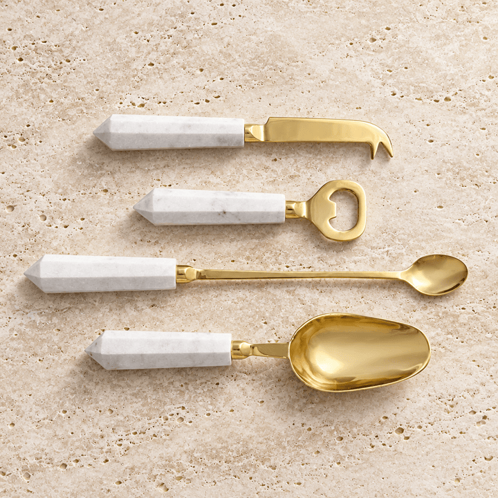Four gold cocktail tools with faceted white marble handles displayed on pale travertine.