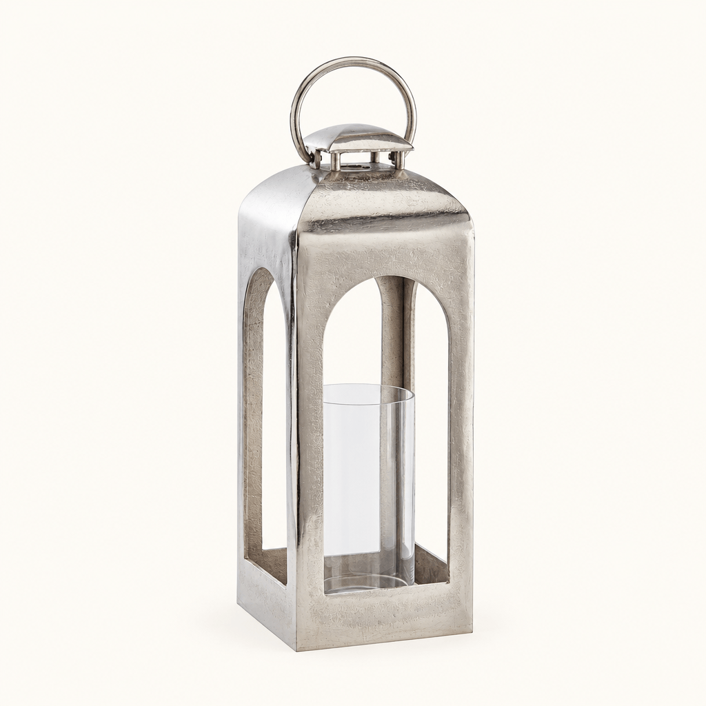 Tall silver aluminum lantern with arched openings, ring handle, and clear glass candle sleeve on an ivory background