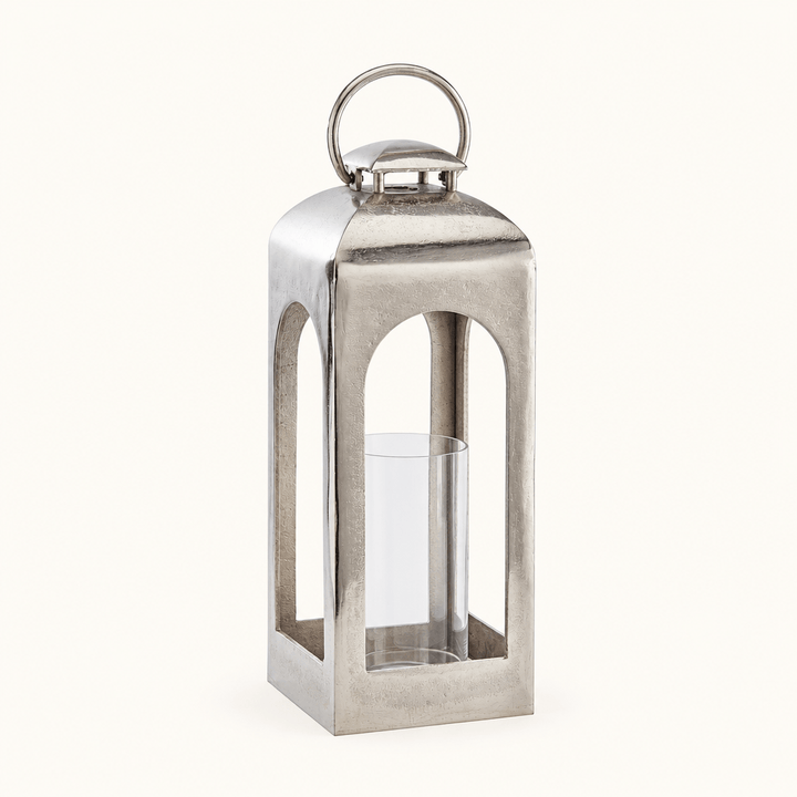 Tall silver aluminum lantern with arched openings, ring handle, and clear glass candle sleeve on an ivory background