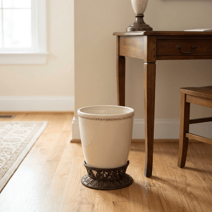 Bellavita cream stoneware wastebasket with ornate dark metal base on a wood floor beside a writing desk