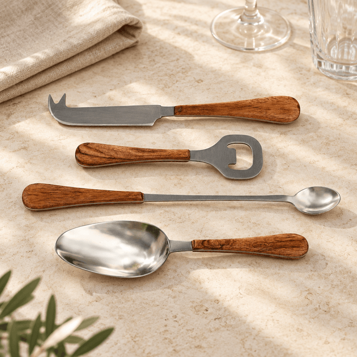 Four acacia wood handled cocktail accessories styled on a cream stone counter beside linen and glassware.