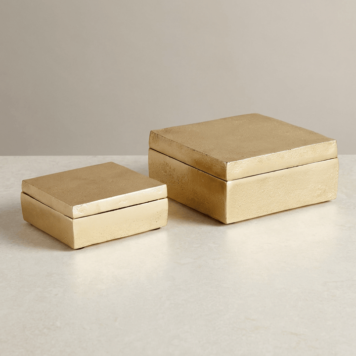 Two gold cast aluminum boxes with flat removable lids displayed on a pale studio surface.