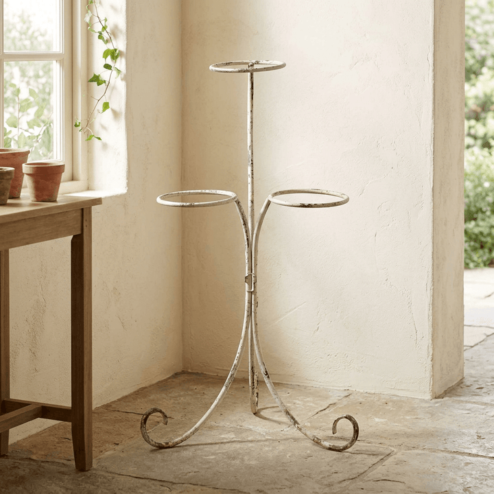 Ivory scrolled iron three-pot plant stand in a sunlit cottage-style room beside a wooden table.