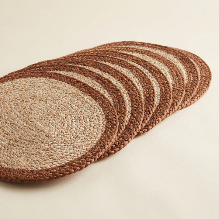 Natural oval jute placemats with rust-brown borders arranged in an overlapping studio display.
