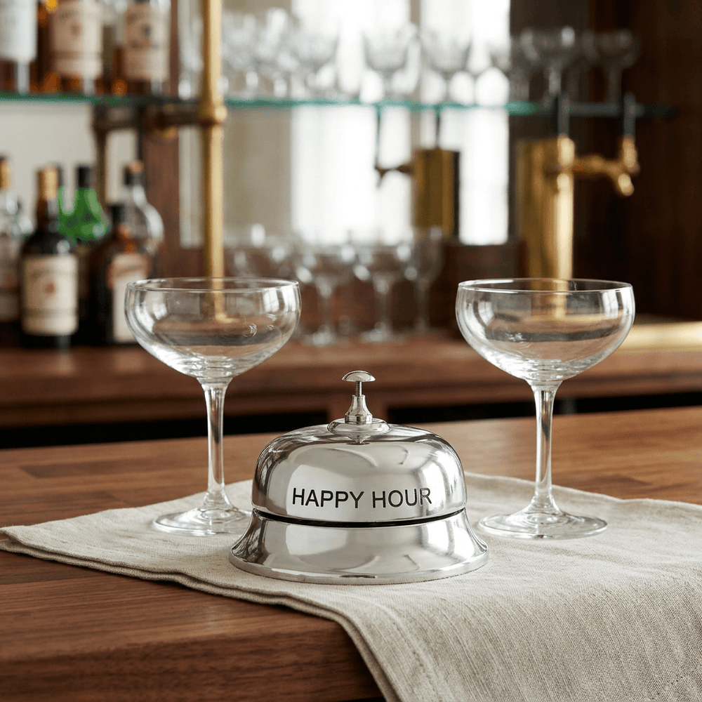 Silver-tone Happy Hour tabletop bell on a wooden bar counter with ivory linen and two coupe glasses.