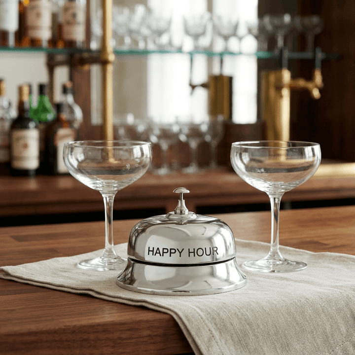 Silver-tone Happy Hour tabletop bell on a wooden bar counter with ivory linen and two coupe glasses.