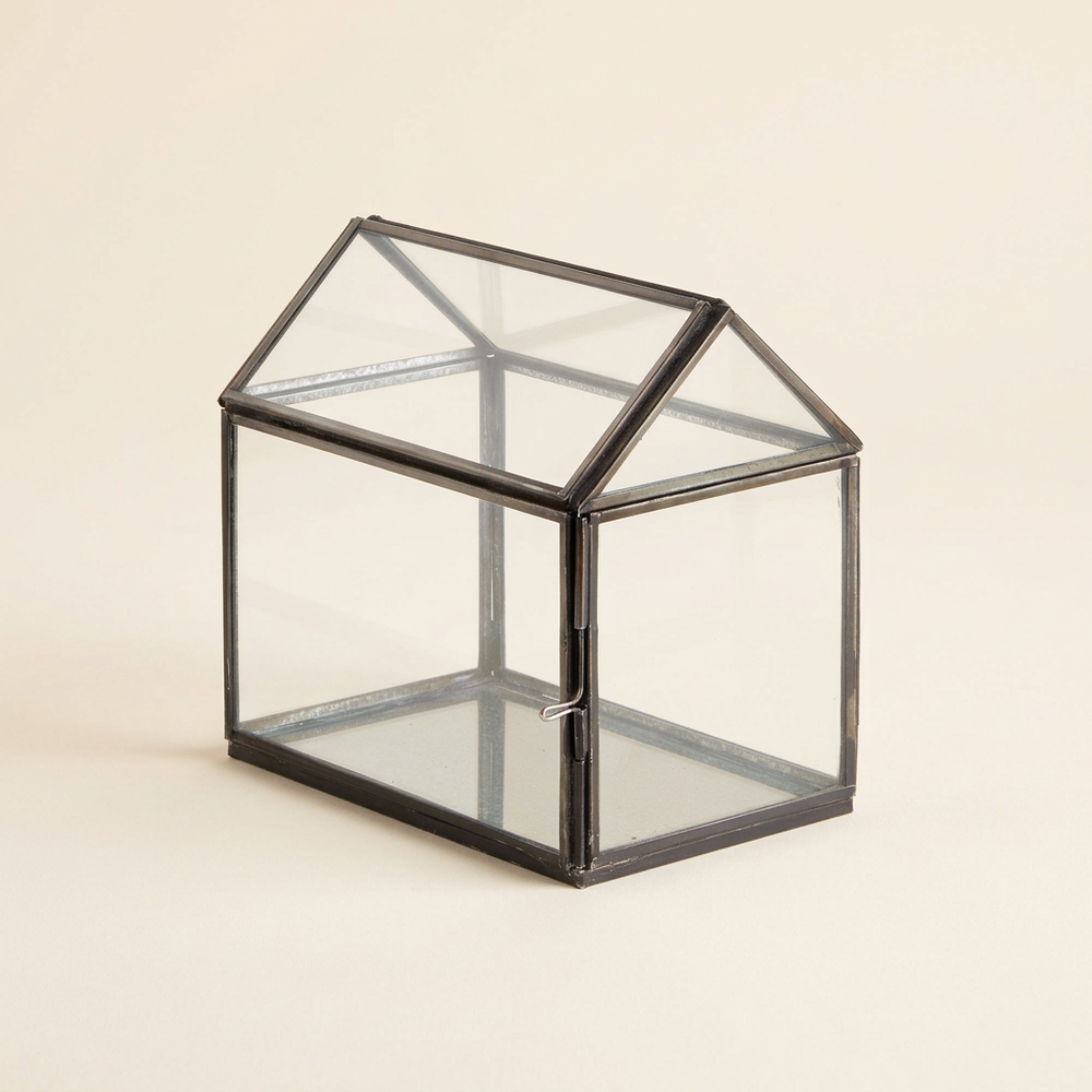 Empty greenhouse-shaped glass terrarium with black iron framing and a pitched roof on an ivory background.