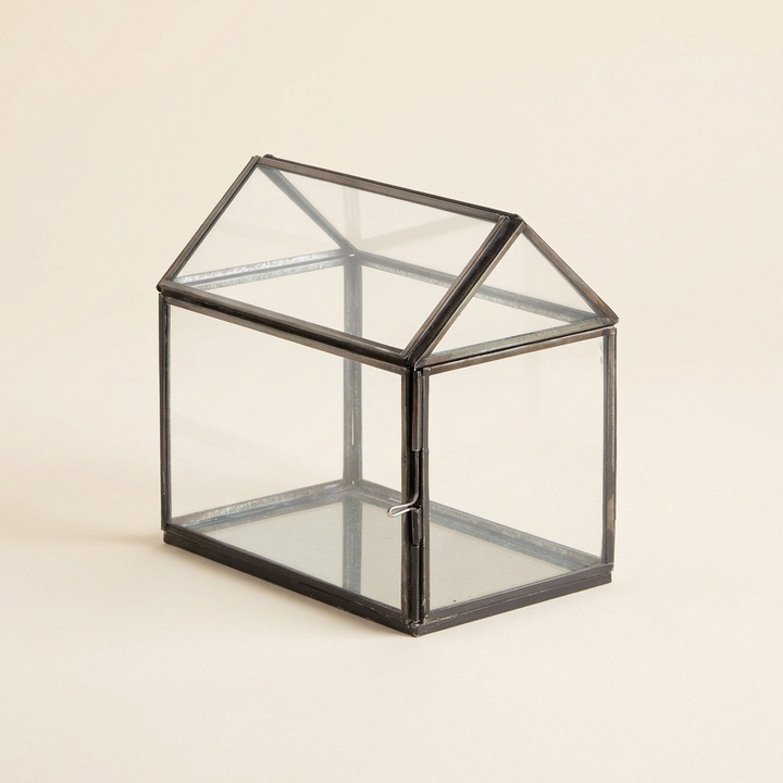 Empty greenhouse-shaped glass terrarium with black iron framing and a pitched roof on an ivory background.