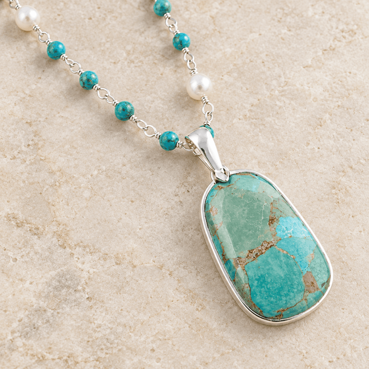 Close-up of the turquoise pendant, sterling silver bezel, and beaded necklace chain on a cream surface
