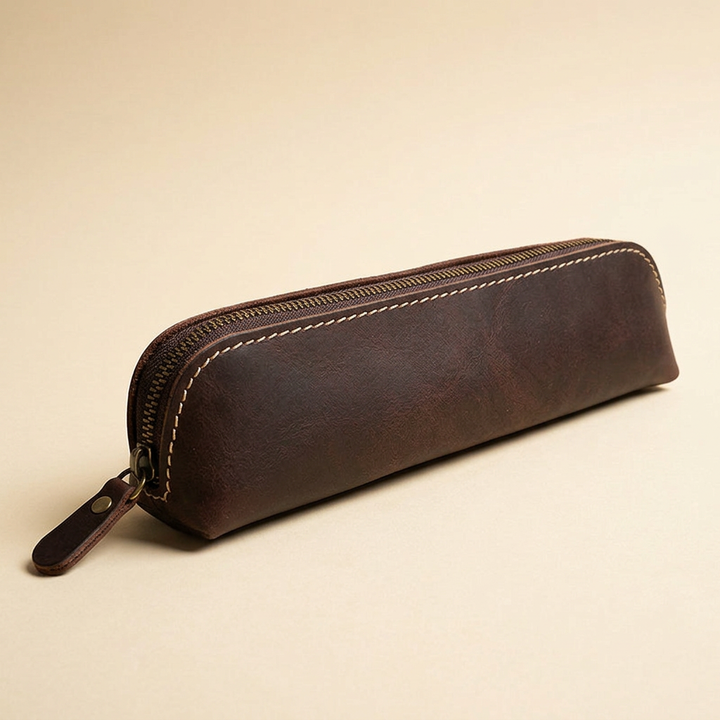 Slim dark brown leather pencil case with metal zipper and leather pull on a warm ivory studio background.