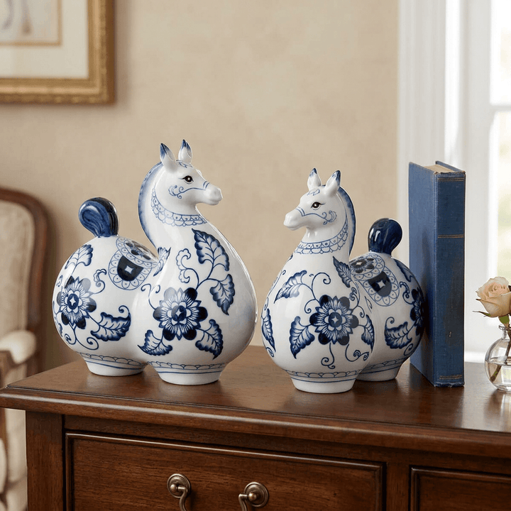 Two blue-and-white floral porcelain horse statues displayed on a wooden console beside a book.