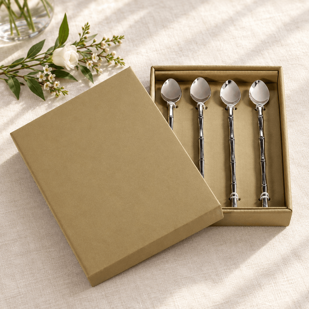 Four silver bamboo-handle stirring spoons presented in their open tan gift box on an ivory linen table.