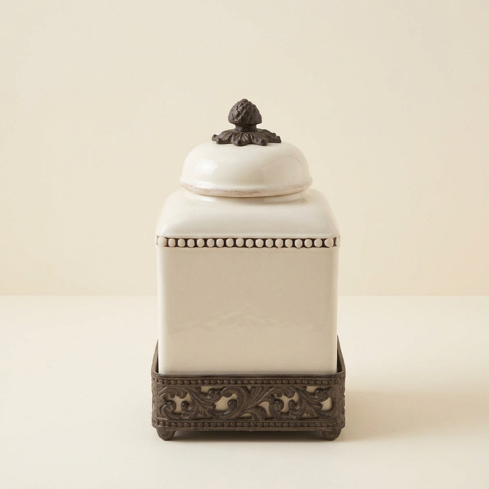 Cream Bellavita square canister with ornate dark metal base and decorative lid on an ivory background.