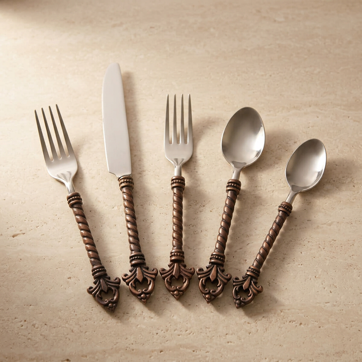 Five Bellavita flatware utensils fanned on a light stone surface, showing twisted handles and ornamental ends.