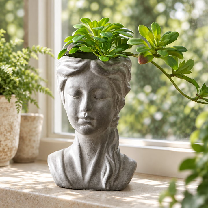 Gray cement girl bust planter with braided hair and succulent foliage on a pale stone windowsill.