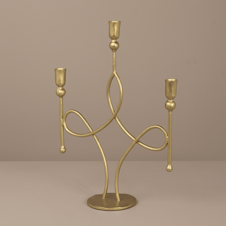 Three-arm antique gold candelabra with looping iron scrolls and a round base against a taupe studio background.