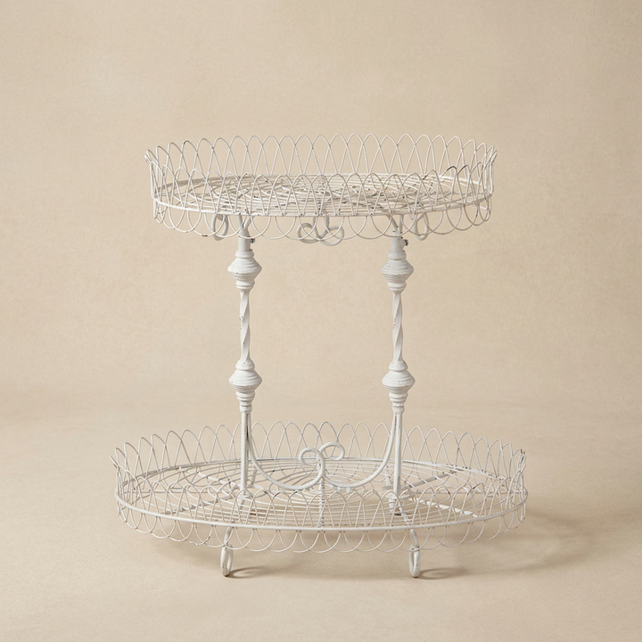 Fleury antiqued white iron plant stand with two oval wire shelves and decorative spindle supports against a warm beige background.