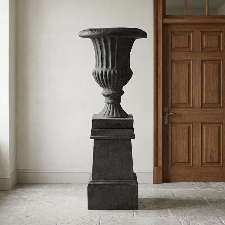 Ashbourne Fluted Urn Planter with Pedestal in Black, shown full length beside a wooden door in a traditional entry hall.
