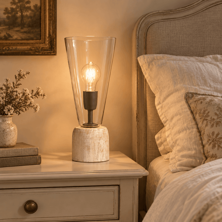 Clear glass and whitewashed wood mini lamp glowing on a cream bedside table beside linen bedding