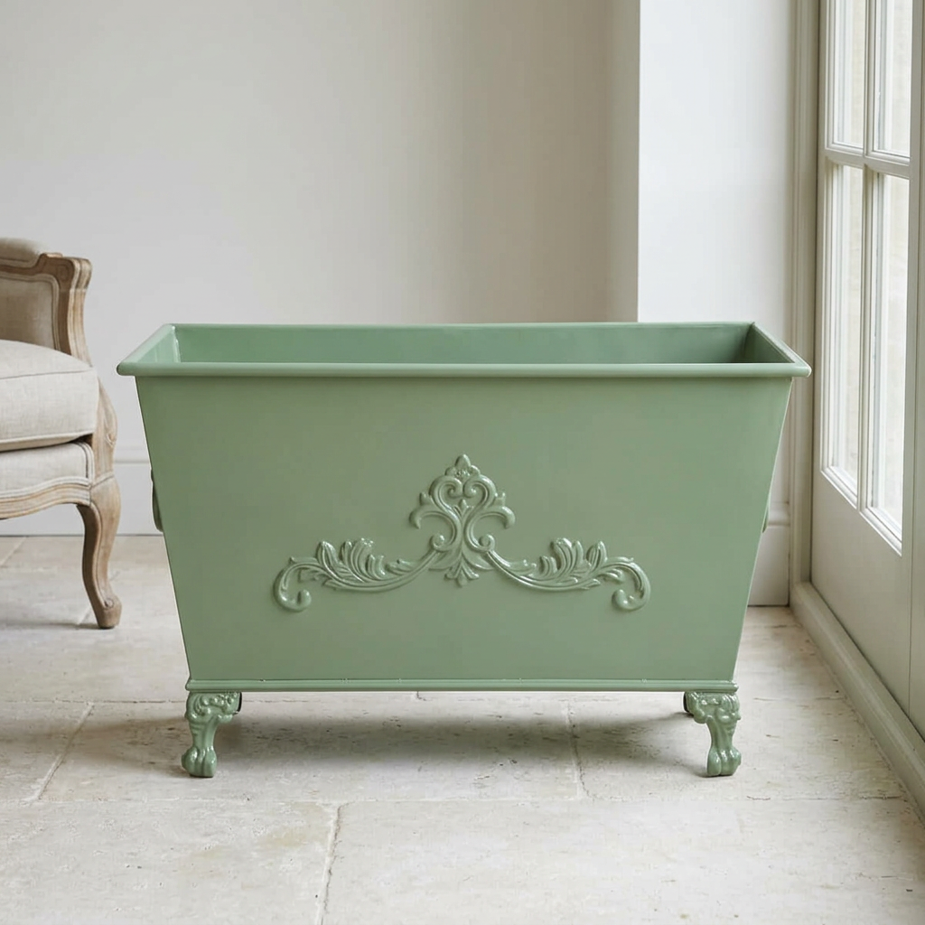 Aveline Scrollwork Metal Planter in Soft Garden Green with raised scrollwork and ornate feet beside a sunroom window.
