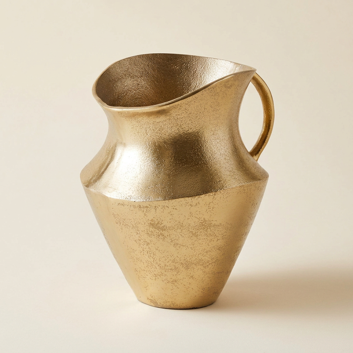 Gold cast aluminum decorative pitcher with an asymmetrical rim and curved handle on an ivory background.