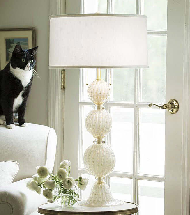 White table lamp with decorative base, black and white cat on a white armchair, and flowers in the foreground.