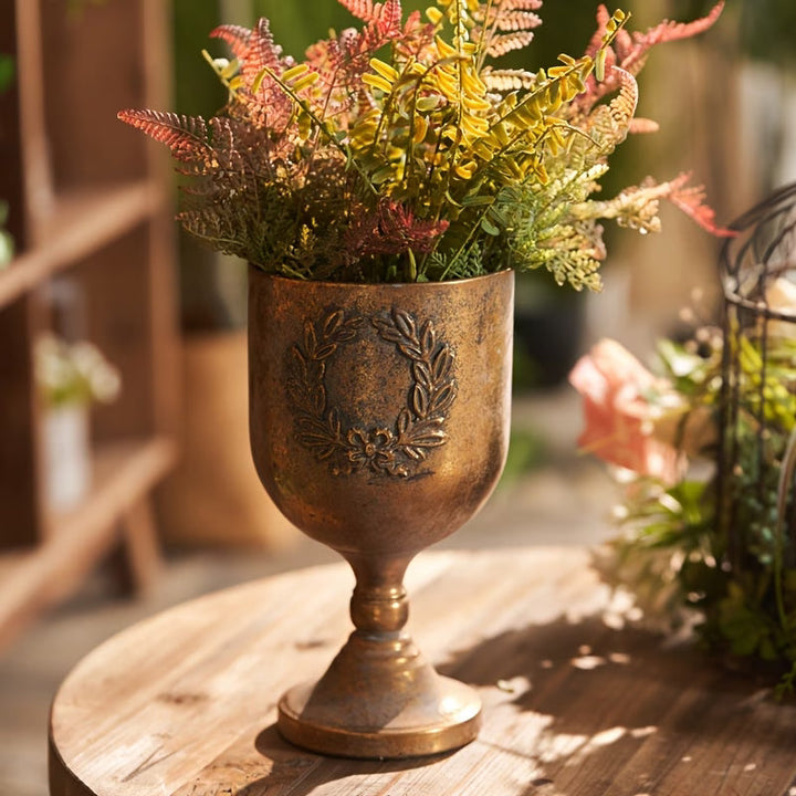 Laurel Wreath Gold Urn Planter filled with greenery on a sunlit wood table