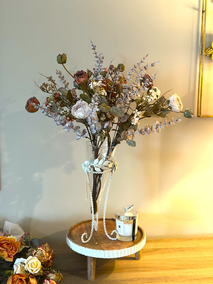 Muted botanical faux flower bouquet in a clear vase beside a candle and pearls
