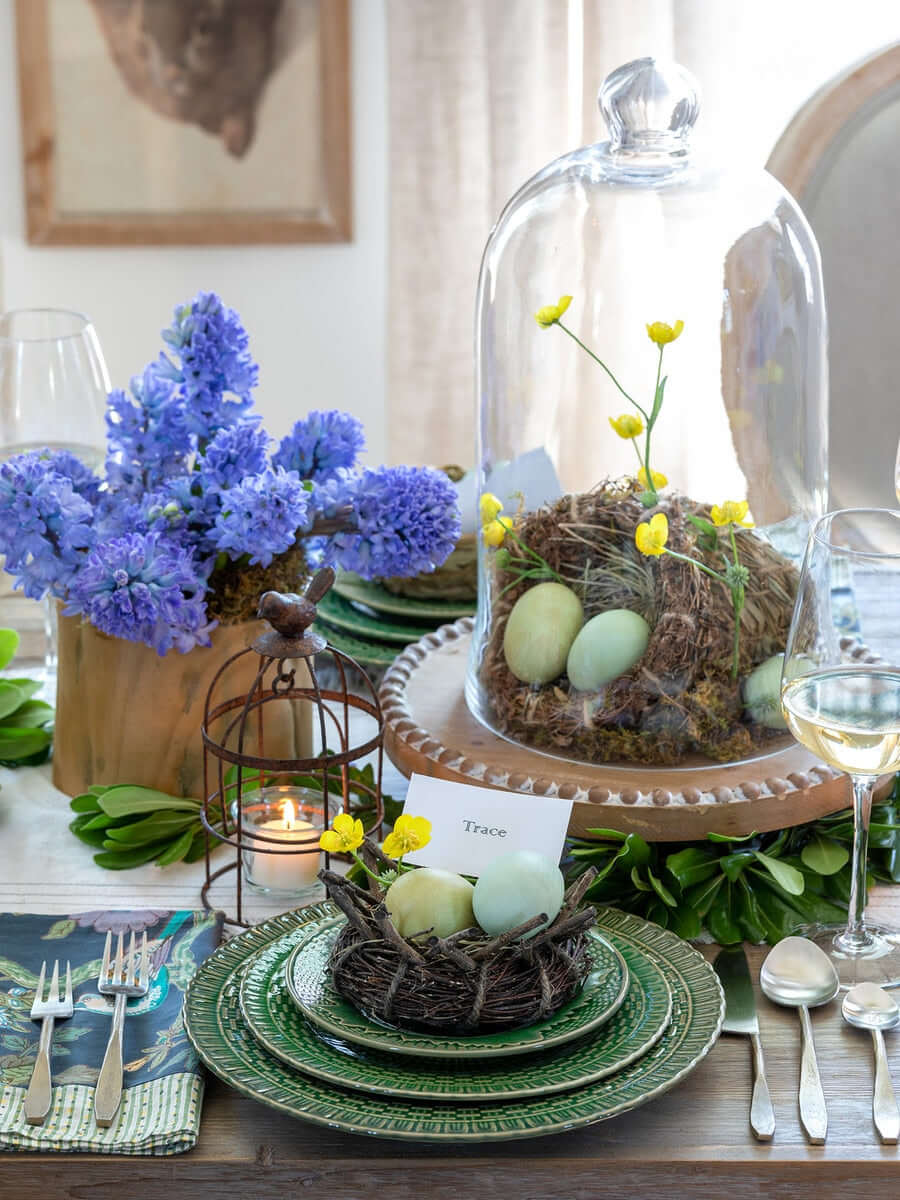 Elegant table setting with natural rattan, grass, and bamboo bird nests, blue flowers, and decorative eggs under a glass cloche.