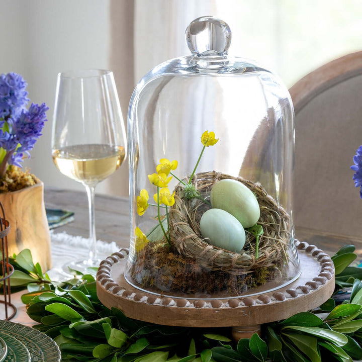 Decorative bird nest with artificial eggs and flowers under a glass cloche on a table with wine and flowers