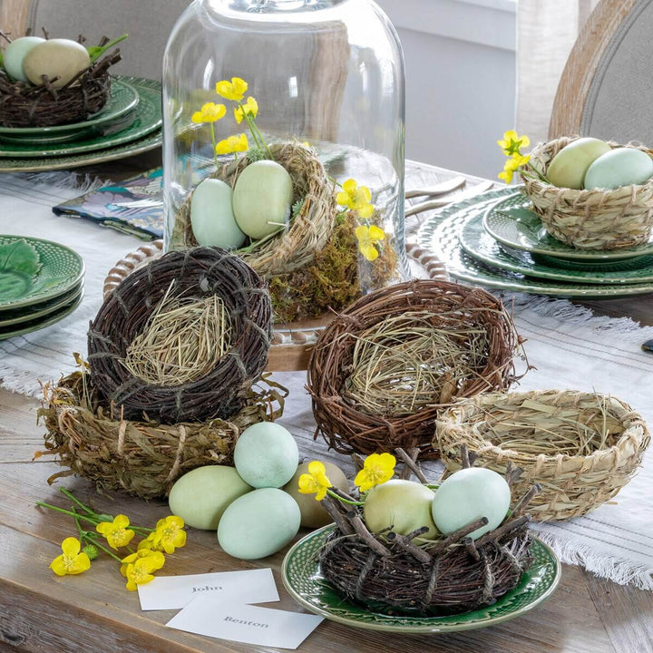 Rustic natural rattan, grass, and bamboo bird nests with decorative eggs and yellow flowers on a dining table.