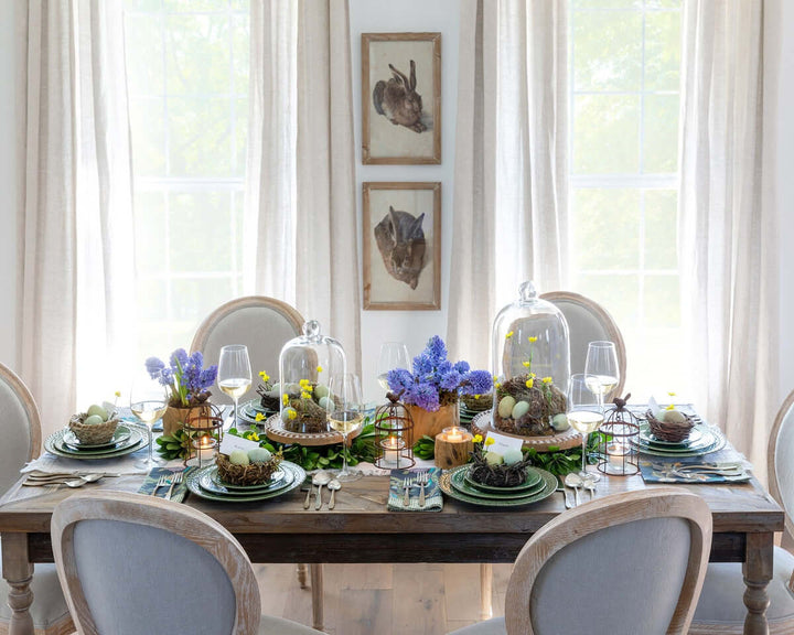 Elegant table setting with natural rattan, grass, and bamboo bird nests, purple flowers, and cloche domes, evoking outdoor beauty.