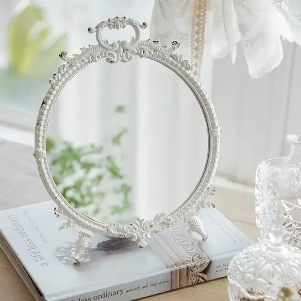 Distressed white vintage makeup mirror with ornate pewter frame on table.