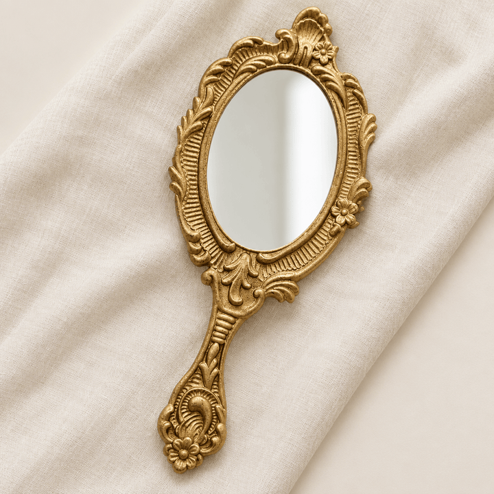 Ornate gold-tone oval handheld mirror styled on ivory linen in a studio setting