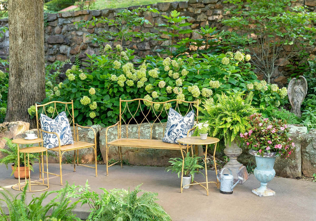 Charming garden scene with ornate metal bench, colorful cushions, potted plants, and lush greenery, creating a picturesque outdoor space.