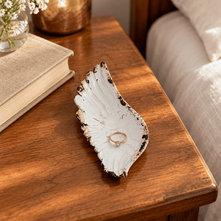 Rustic white pewter angel wing jewelry tray holding a small ring beside a book on a wooden bedside table
