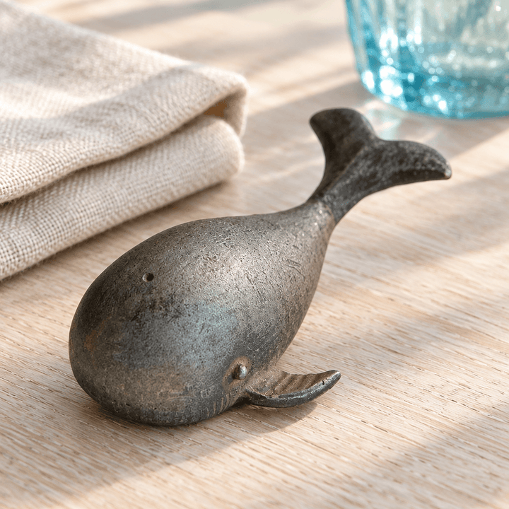 Pewter whale bottle opener on a pale oak table with natural linen and a blue drinking glass.