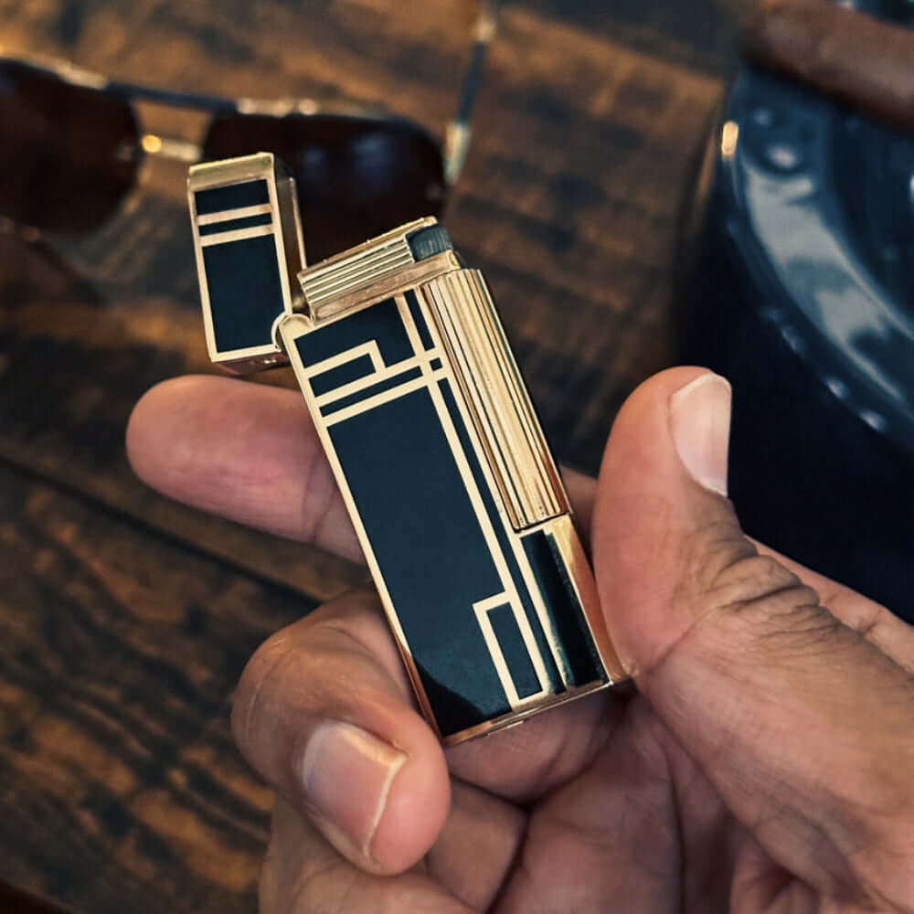 The Prestige Art Deco Lighter in Black and Gold held by a hand on a wooden table.