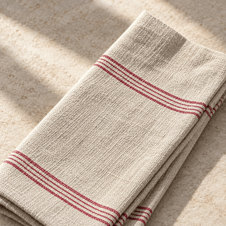 Close-up of beige woven cotton napkin showing fine dark red stripes and a finished hem.