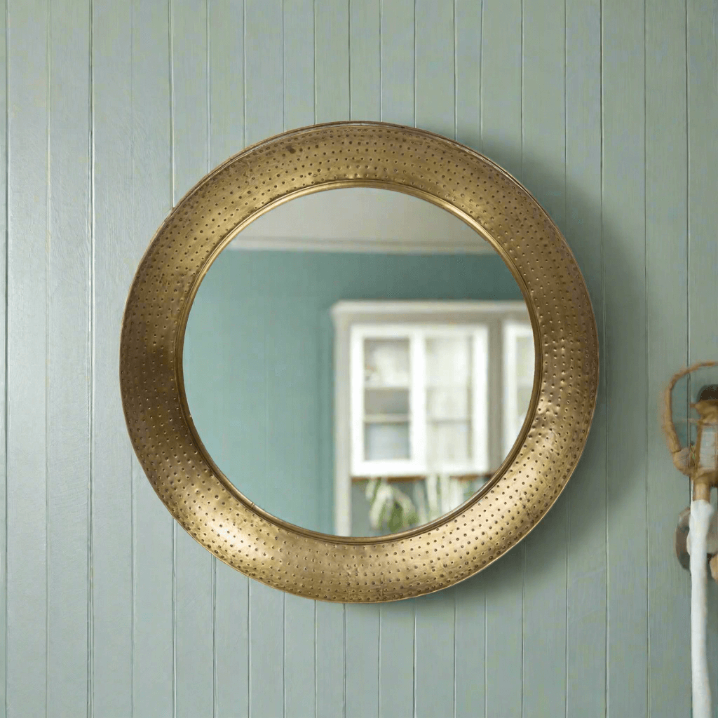 Round hammered brass wall mirror with vintage iron porthole design, reflecting a serene indoor space. Elegant and timeless home decor.
