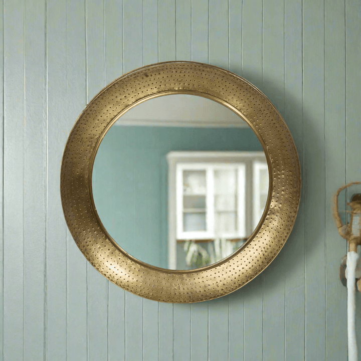 Round hammered brass wall mirror with vintage iron porthole design, reflecting a serene indoor space. Elegant and timeless home decor.
