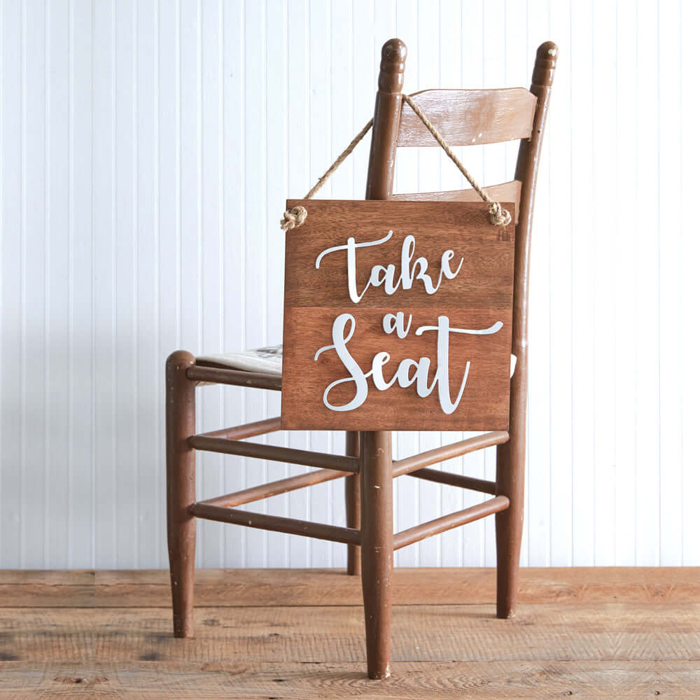 Rustic wood 'Take a Seat' sign with white lettering and jute rope hanging on a wooden chair, perfect for home decor.