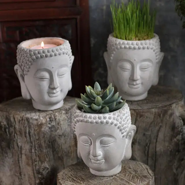 Three cement Buddha head planters displayed on wood stands with a succulent, grass and a candle; each vessel sold separately