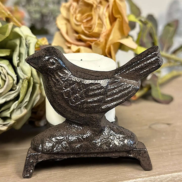 Dark sculpted metal bird candle stand holding a small white candle among muted flowers